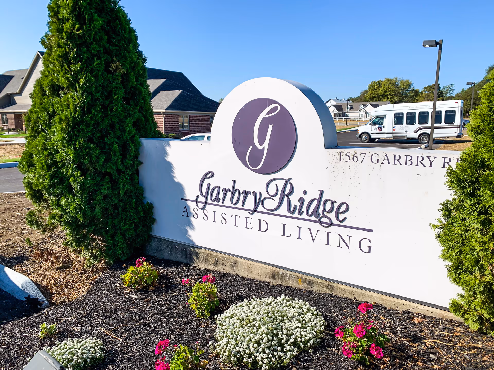 Outdoor view of a white sign with purple lettering that reads 'Garbry Ridge Assisted Living' surrounded by landscaped flowers and greenery, with a building and a white shuttle bus in the background under a clear blue sky.