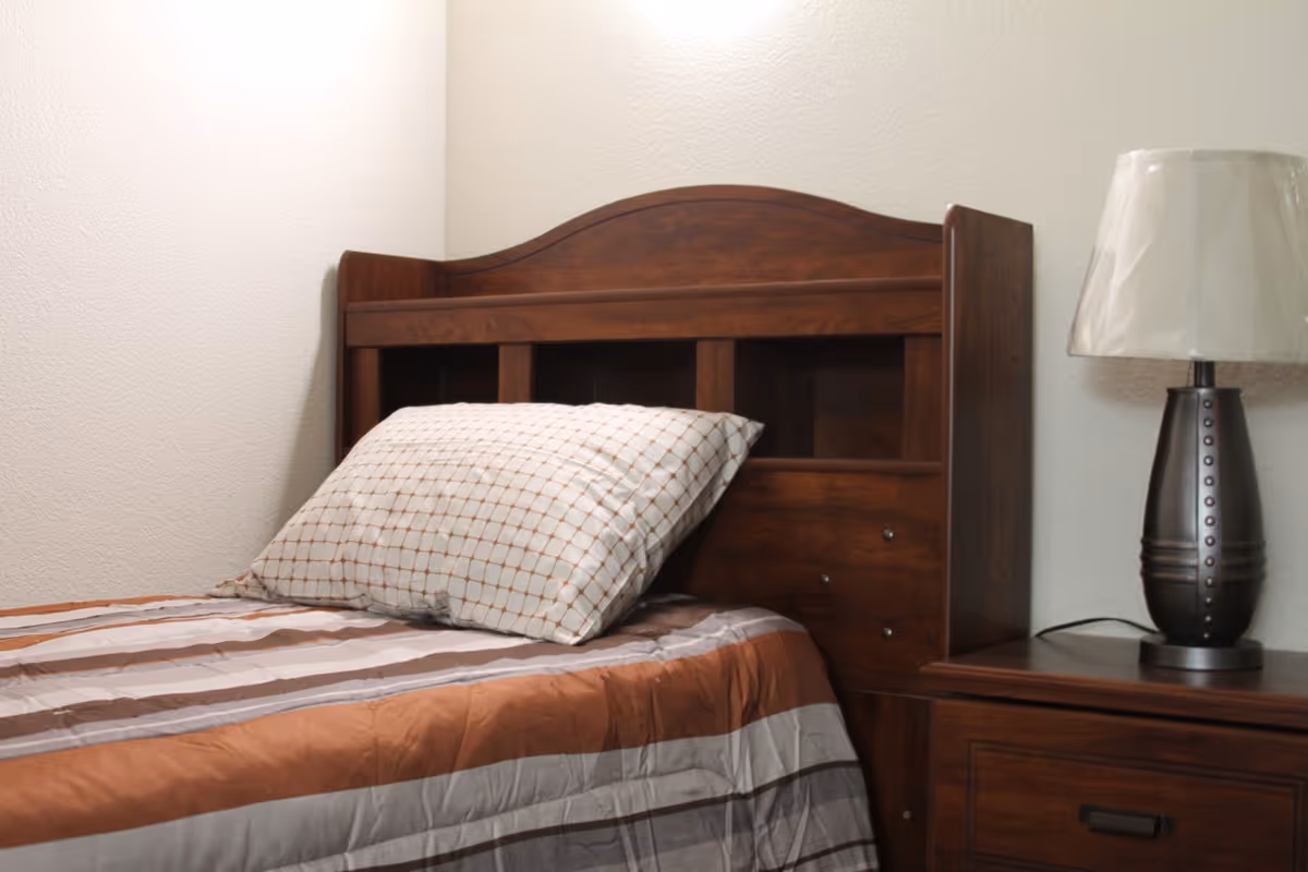A neatly made single bed with a wooden headboard and a pillow with a checkered pattern. Next to the bed is a wooden nightstand with a dark-colored lamp that has a white lampshade.