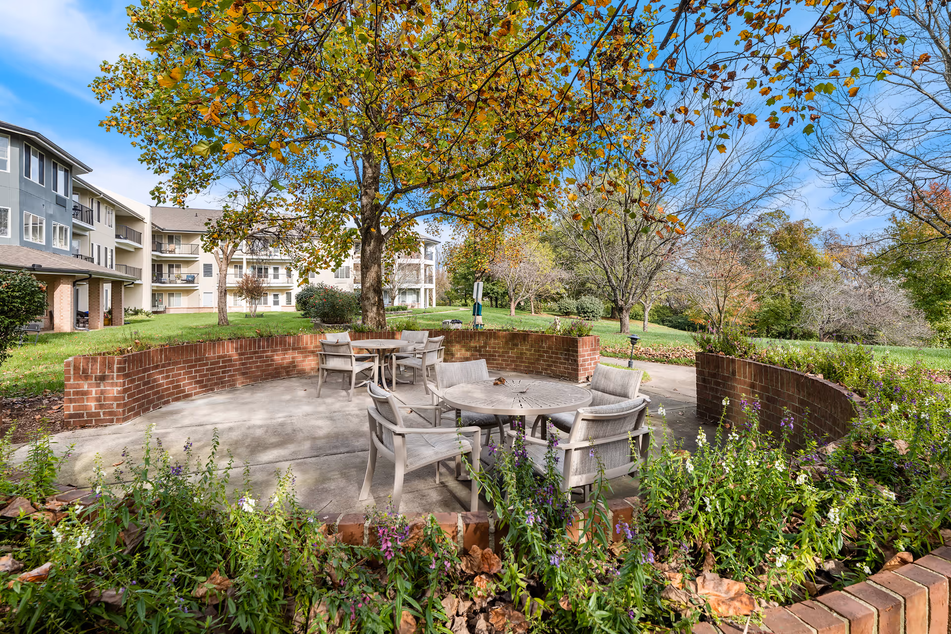 Outdoor seating area with round tables and chairs surrounded by a low brick wall, trees with autumn leaves, and a grassy lawn with senior living community buildings in the background under a blue sky.