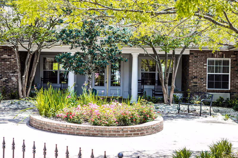 Front entrance of a brick building with a circular flower bed, trees, benches, and a porch with rocking chairs.