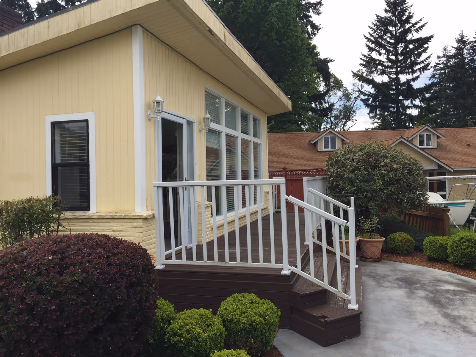 Exterior view of a yellow single-story building with large windows and a small wooden deck with white railings. The deck has steps leading down to a concrete pathway surrounded by neatly trimmed bushes and plants. In the background, there is another building with a brown roof and dormer windows, along with tall trees.