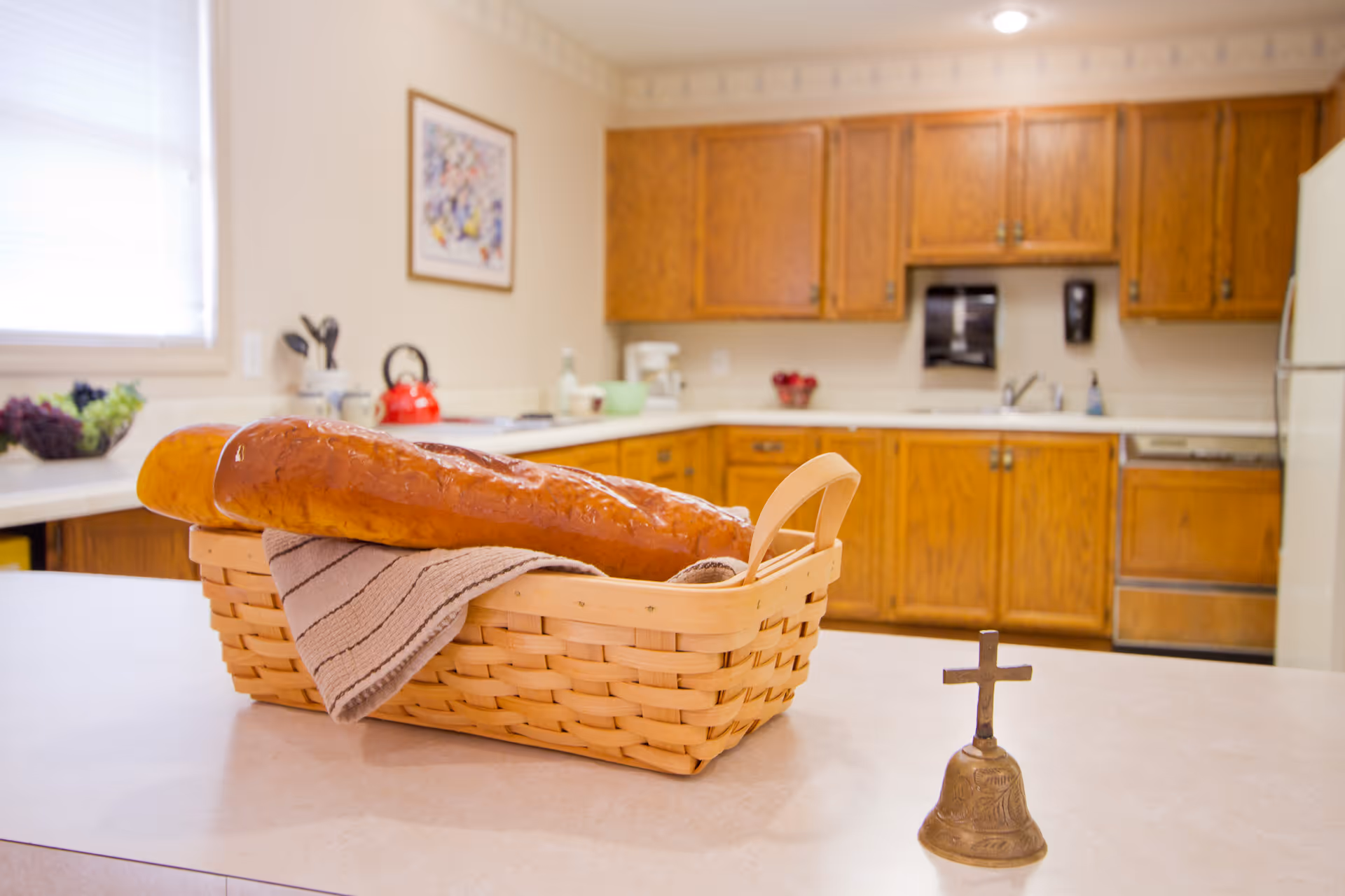 A kitchen interior with wooden cabinets and white countertops. On the countertop in the foreground, there is a woven basket containing two baguettes and a striped cloth. Next to the basket is a small brass bell with a cross on top. In the background, there is a red kettle, a coffee maker, and a framed picture on the wall.