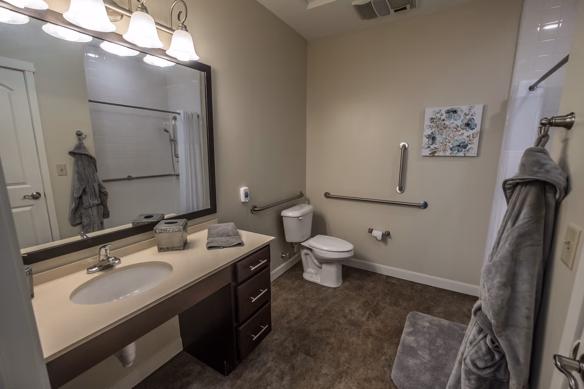 A clean, accessible bathroom with a large mirror above a sink and countertop. The bathroom features a toilet with grab bars on the walls for support, a towel rack with a gray robe hanging, a gray bath mat on the floor, and a shower with a white curtain. There is a decorative floral picture on the wall.