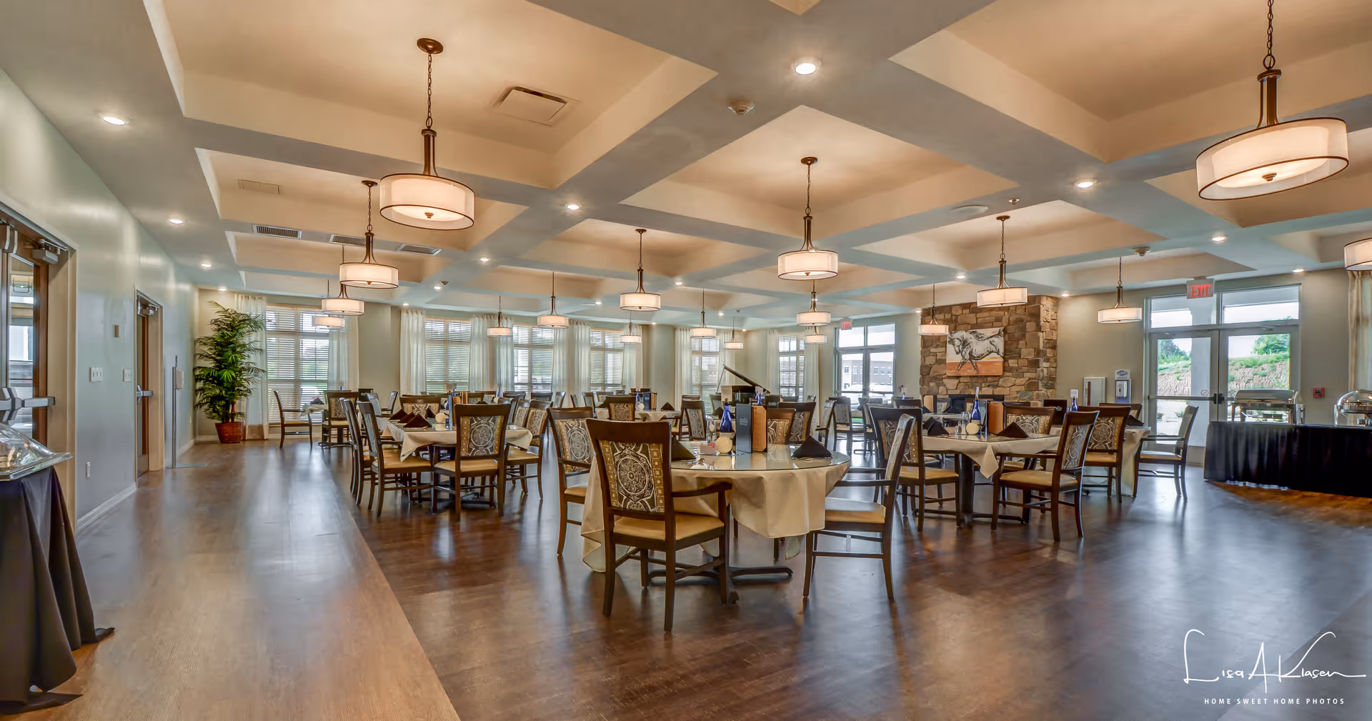 A spacious dining room with multiple tables covered with beige tablecloths and set with blue water bottles and napkins. The room features wooden chairs with patterned cushions, large windows allowing natural light, modern ceiling lights, and a stone accent wall with a horse painting. The floor is wooden and there are plants and doors visible along the walls.