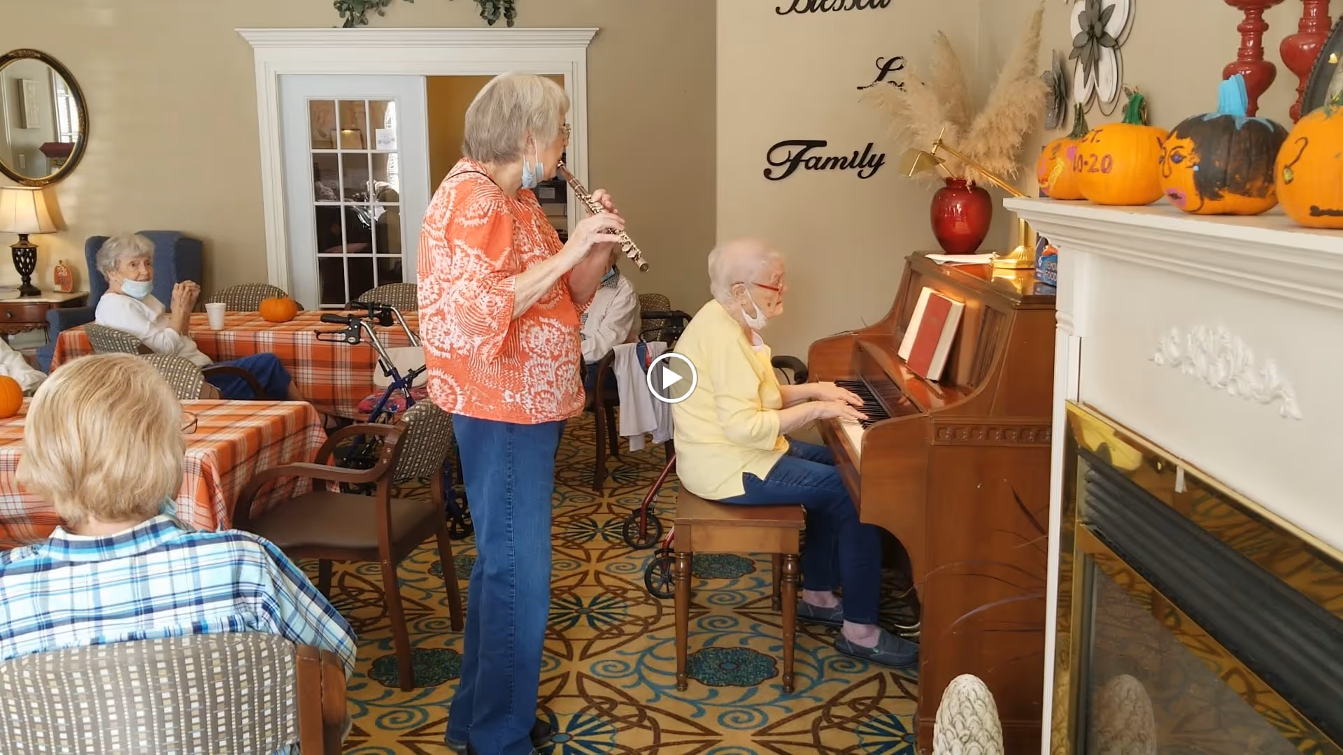 Seniors gathered in a common room where a woman plays the piano and another plays a flute while others sit at tables.