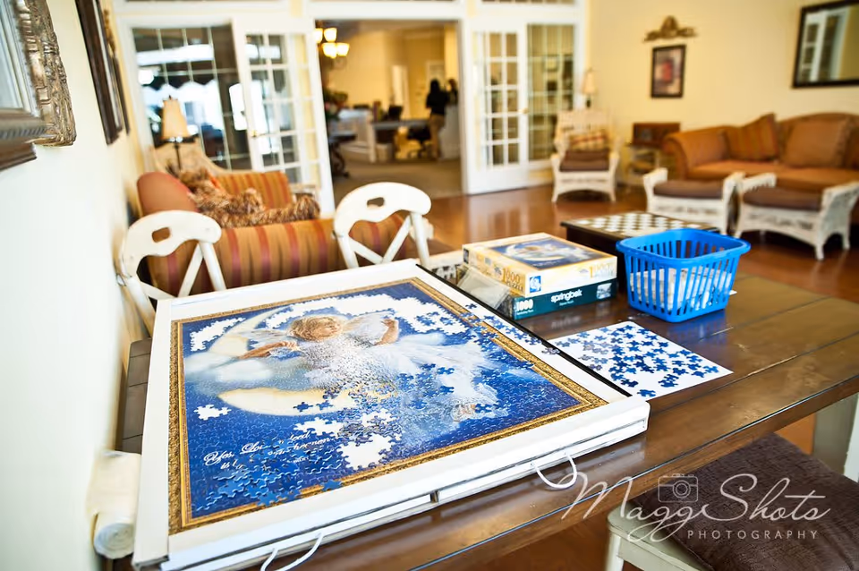 A cozy living room area with a wooden table in the foreground featuring a partially completed jigsaw puzzle, puzzle boxes, and a blue basket. The background shows comfortable seating including striped and cushioned chairs and sofas, with a bright and inviting atmosphere.