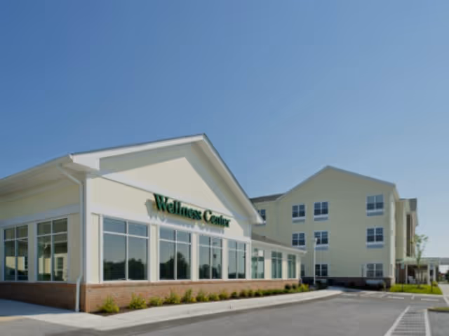 Exterior view of a building labeled Wellness Center with large windows and a clear blue sky above. The building is part of a larger facility with multiple floors and parking spaces in front.