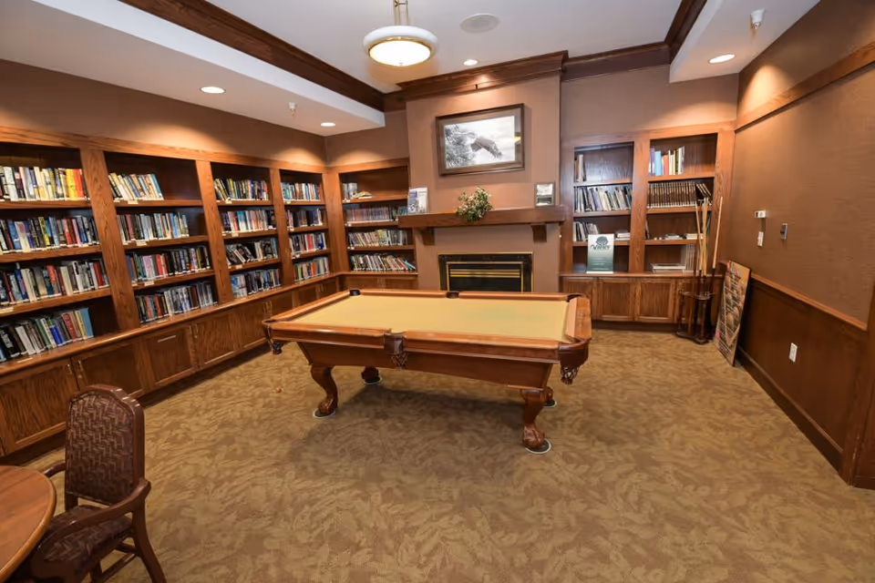 A cozy room with a pool table in the center, surrounded by wooden bookshelves filled with books. There is a fireplace with a framed picture above it on the far wall. The room has warm brown walls and carpeted floor with a leafy pattern. A chair and part of a round table are visible in the foreground.