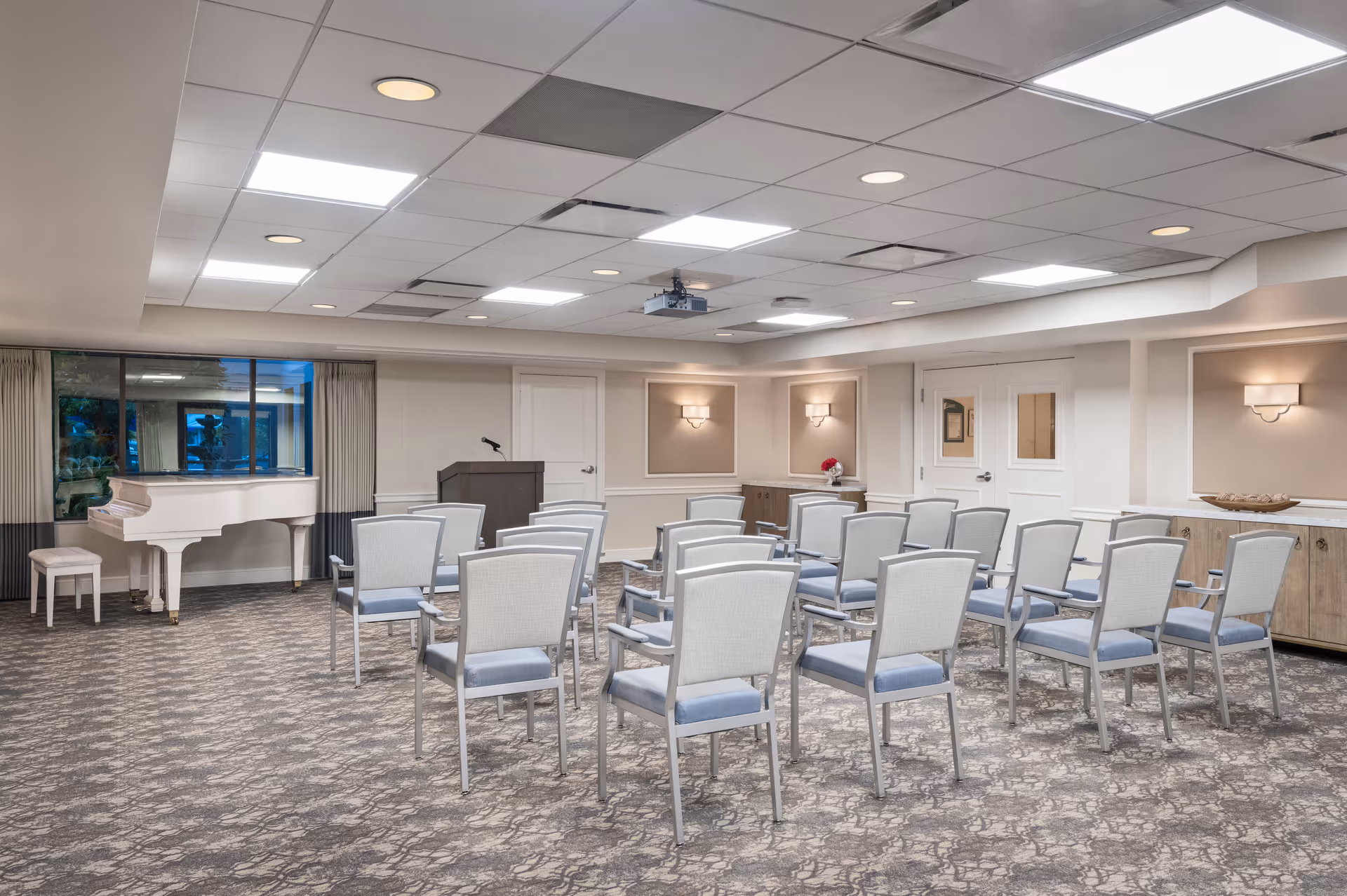 A well-lit meeting or presentation room with rows of gray cushioned chairs facing a wooden podium with a microphone. There is a white grand piano with a matching bench near a window with curtains. The room has a patterned carpet, recessed lighting in the ceiling, and wall sconces providing additional light. Cabinets line the walls, and a projector is mounted on the ceiling.