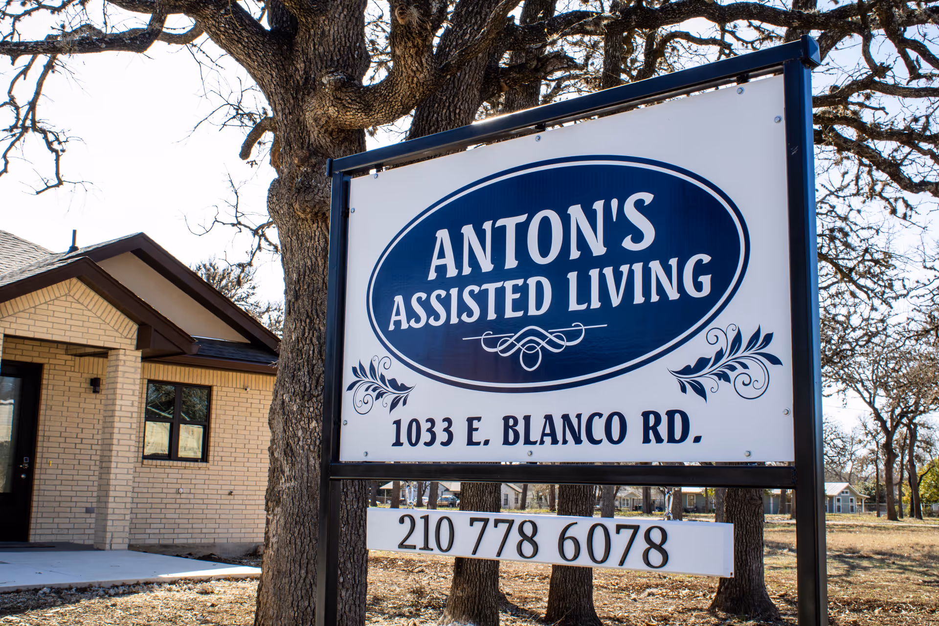 Outdoor sign for Anton's Assisted Living displaying the address and phone number in front of a brick building and trees.