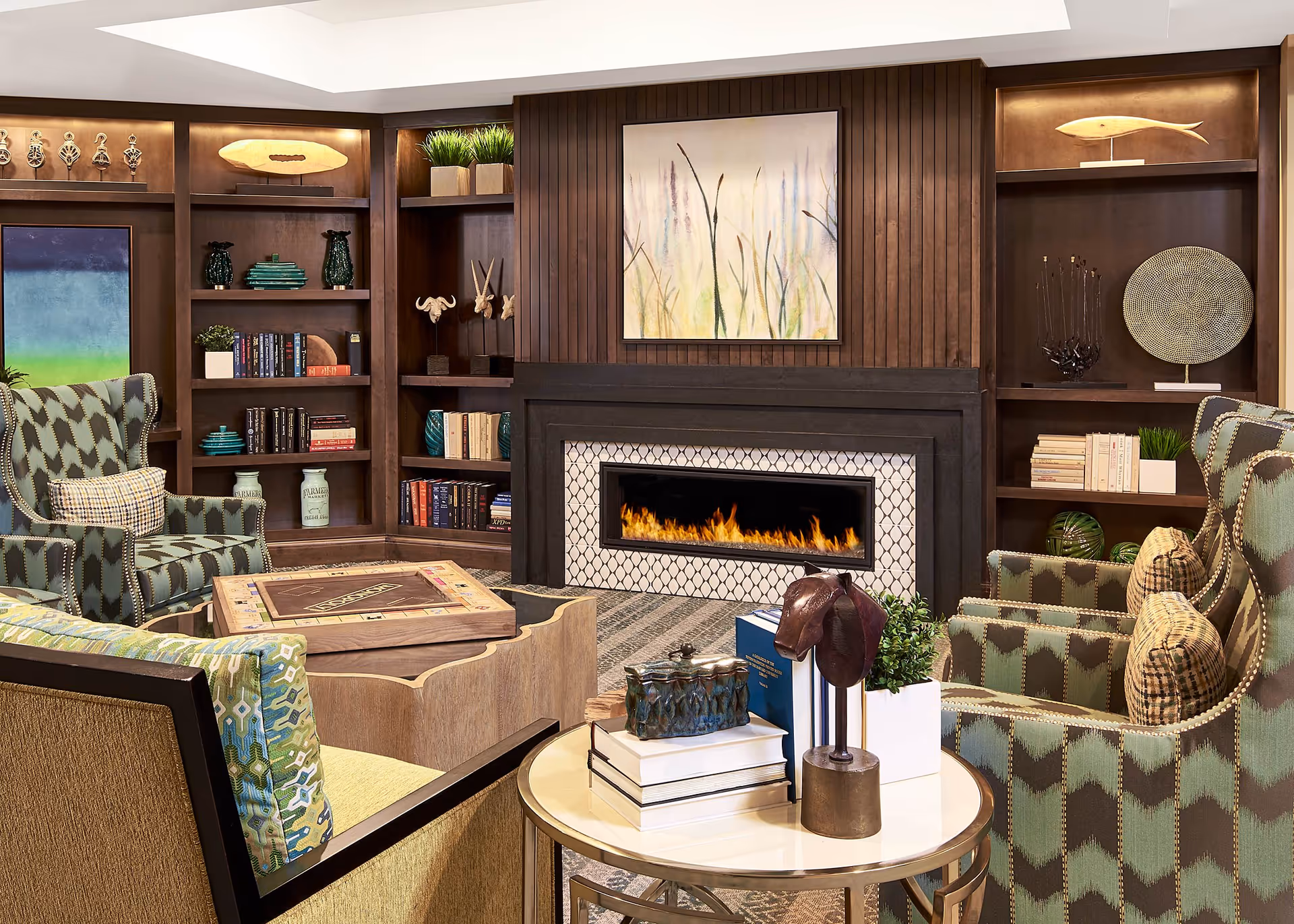 A cozy living room area with patterned armchairs and a sofa arranged around a modern fireplace with a tiled surround. The room features built-in wooden bookshelves filled with books and decorative items, a coffee table with a board game, and a round side table with books and a small sculpture. Above the fireplace hangs a painting of tall grasses.
