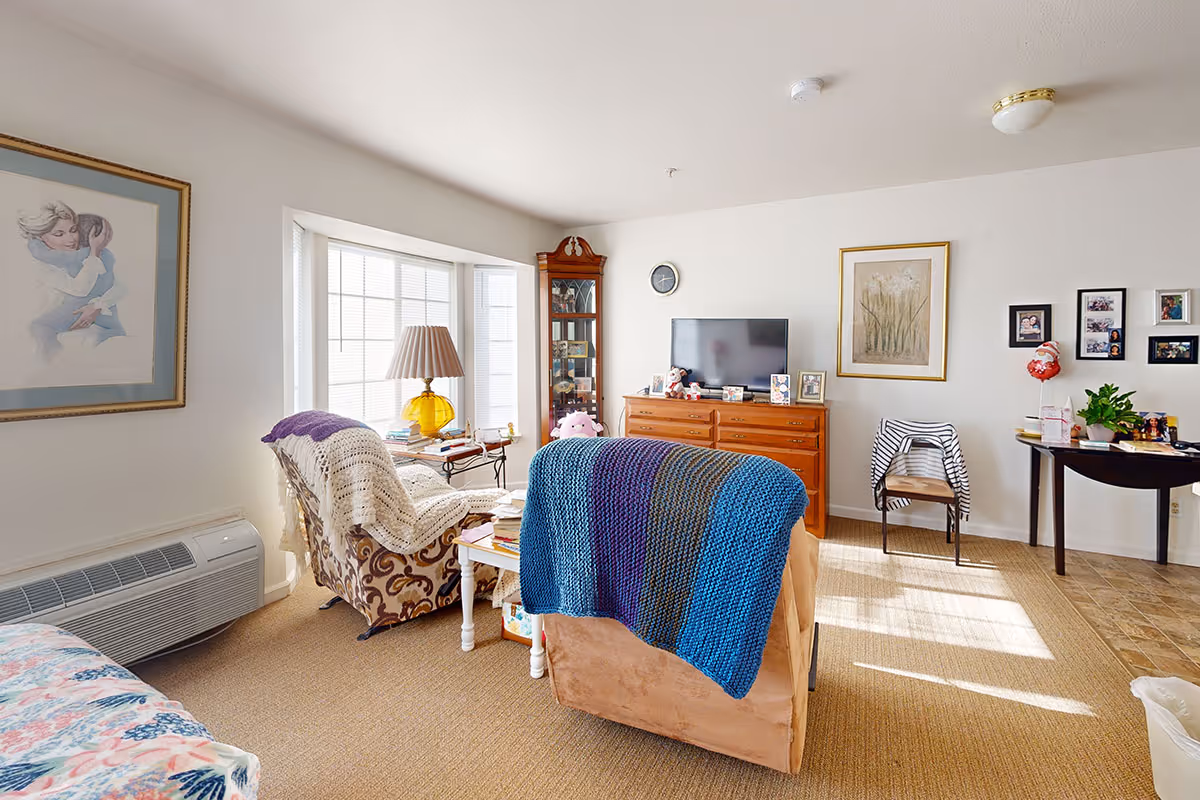 Well-lit senior living room with armchairs, a TV on a dresser, a bay window, and framed artwork.