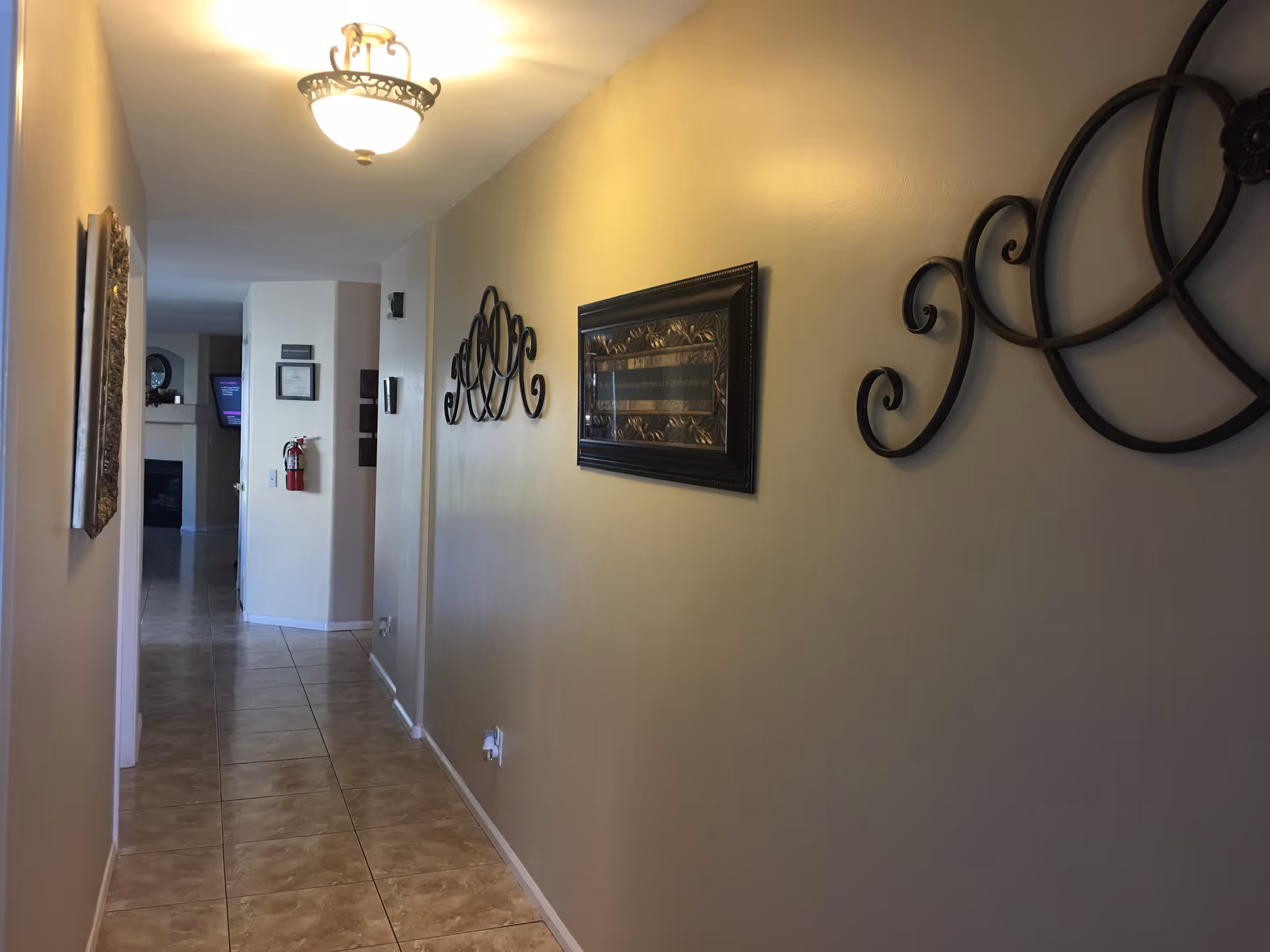 A narrow hallway with beige walls and tiled floor, decorated with black wrought iron wall art and a framed picture. A ceiling light fixture illuminates the space. At the end of the hallway, a fire extinguisher is mounted on the wall and a television is visible in an adjoining room.
