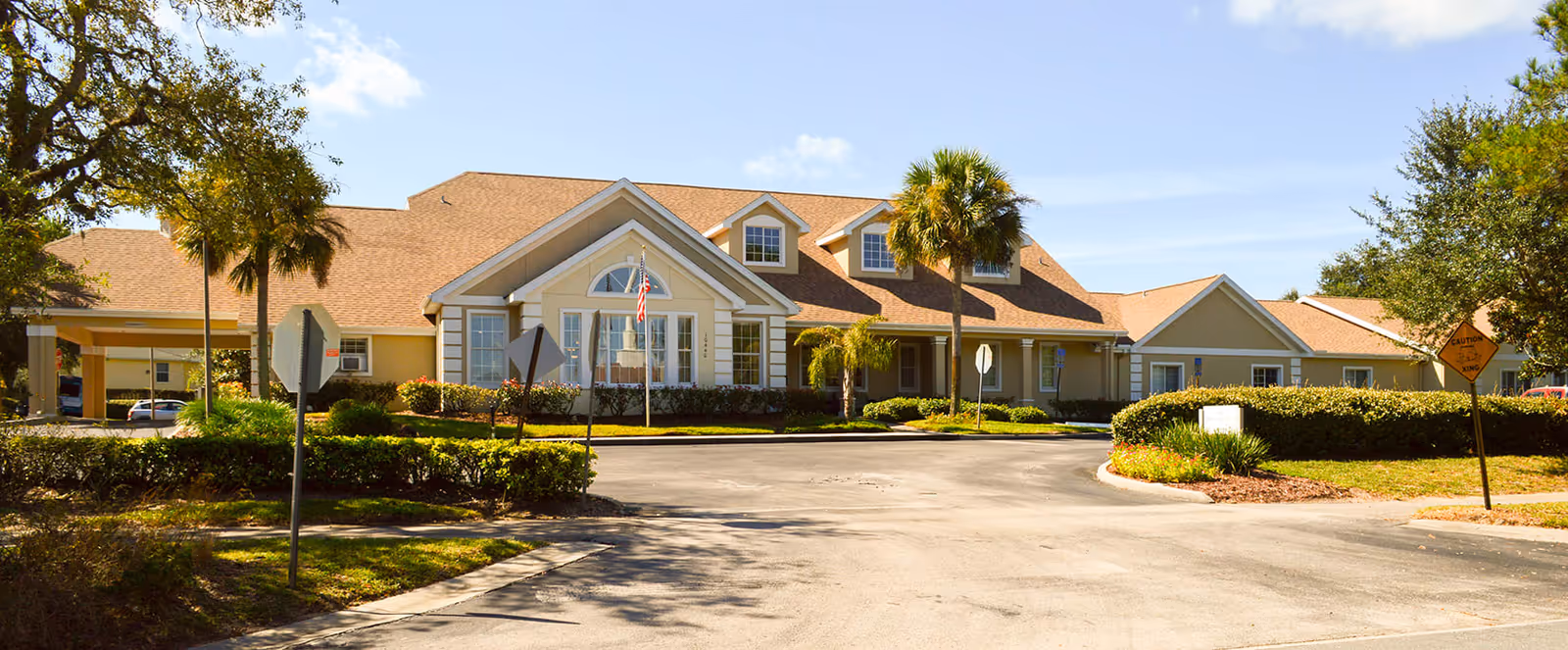 Exterior view of a single-story senior living facility building with a beige facade and brown shingle roof, surrounded by palm trees and shrubs under a clear blue sky.