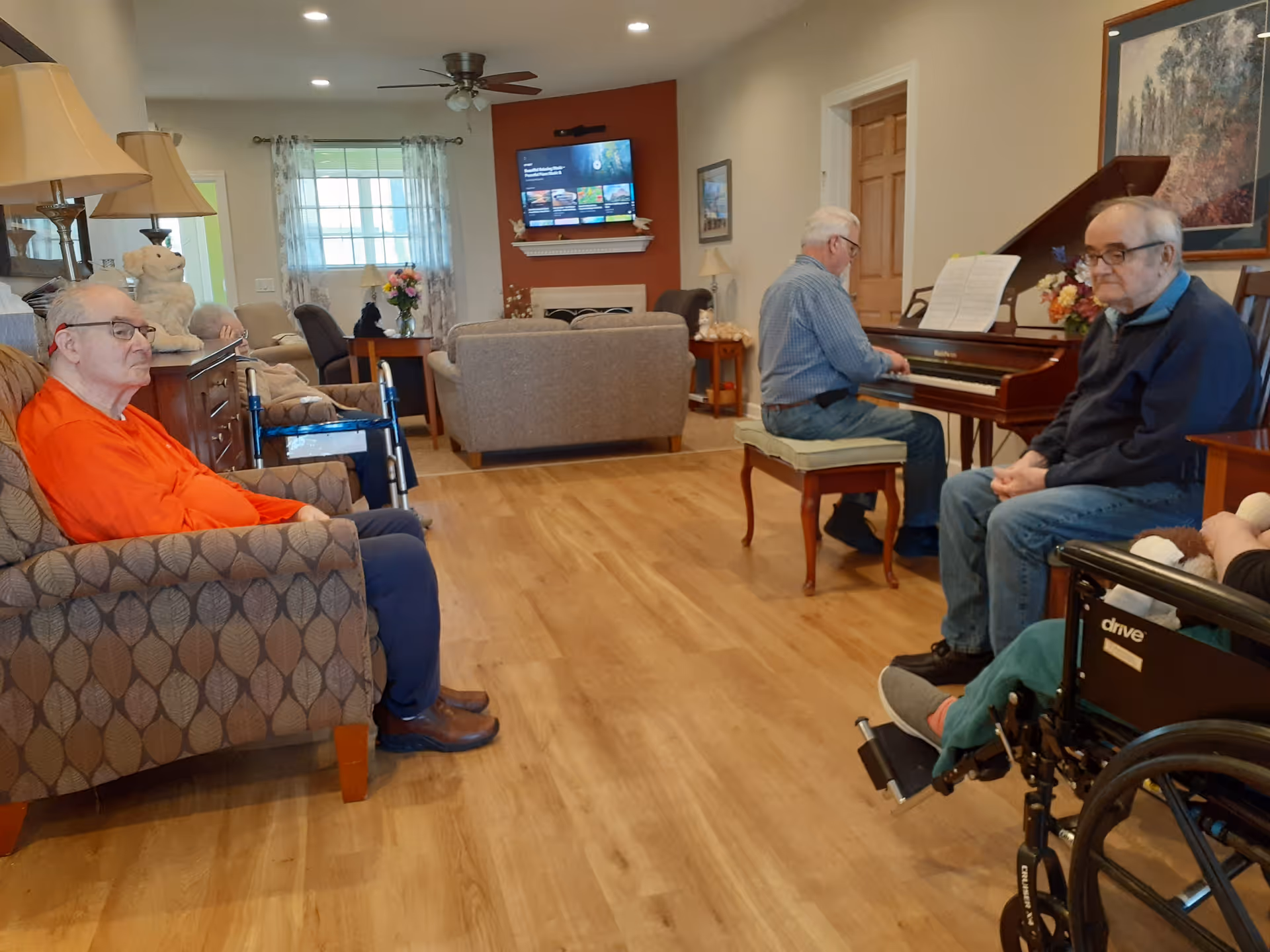 A bright common living room with several residents seated while one person plays a piano, with couches, a TV, and a wheelchair visible.