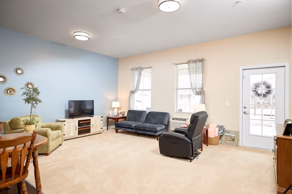 A bright living room with beige carpet and walls painted in light blue and beige. The room features a green armchair, a blue sofa, a black recliner chair, a wooden TV stand with a flat-screen TV, two side tables with lamps, and a wooden dining table with chairs. Two windows with sheer curtains and a glass door with a wreath allow natural light into the space.