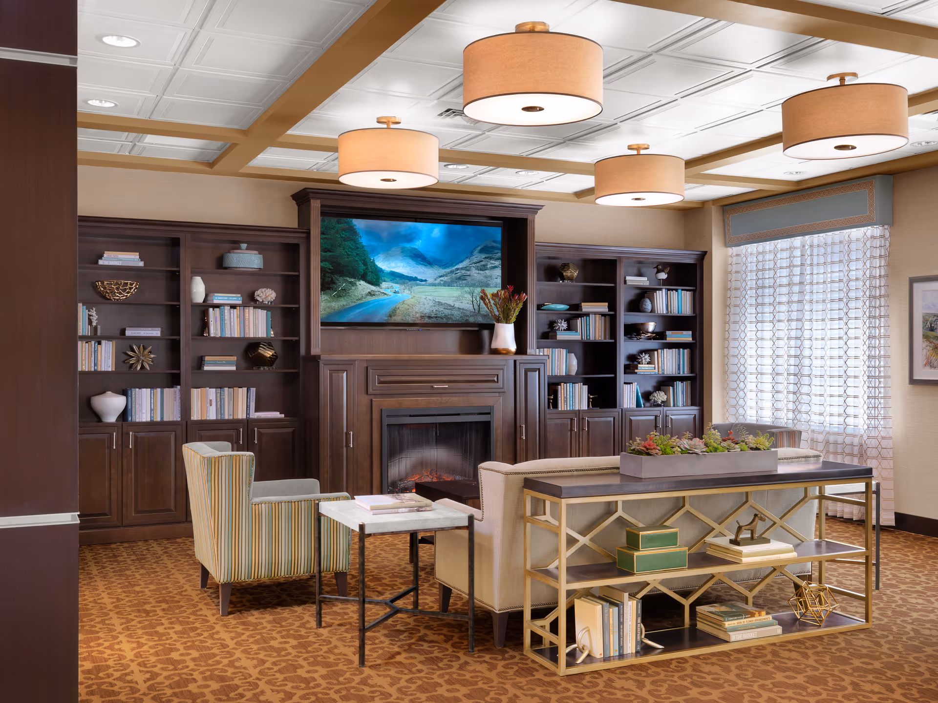 A cozy senior living facility lounge area with a beige sofa, a striped armchair, a small side table, and a decorative console table with books and ornaments. The room features built-in dark wood bookshelves filled with books and decorative items, a fireplace with a large flat-screen TV above it displaying a scenic landscape, patterned carpet, and large windows with sheer curtains allowing natural light.