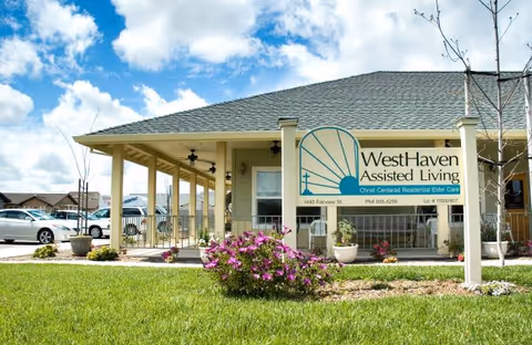 Exterior view of Westhaven Assisted Living facility showing a single-story building with a covered porch, a sign with the facility name and contact information, green lawn, and flowering bushes under a partly cloudy sky.