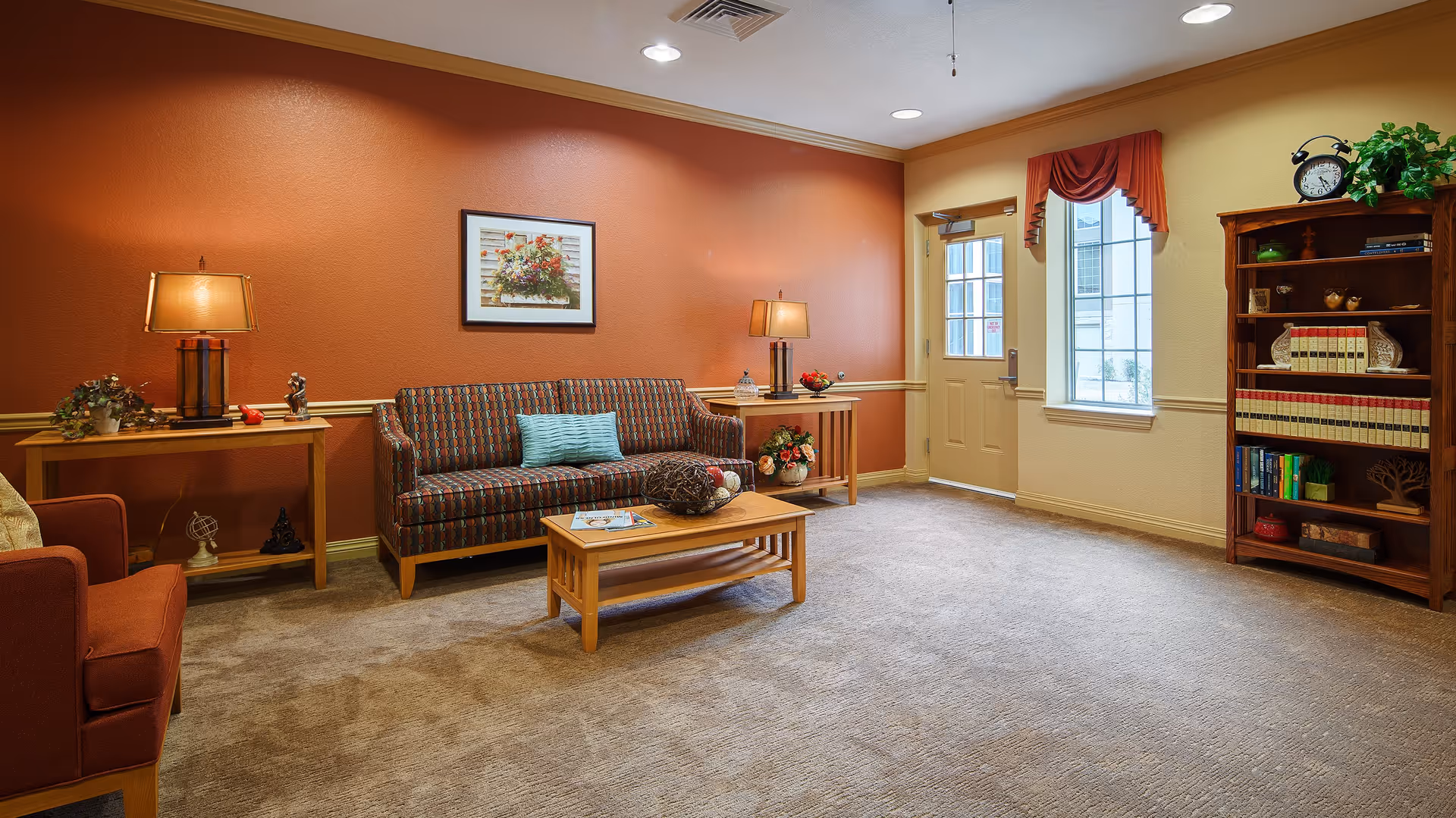 A cozy living room with a patterned sofa, an armchair, two wooden side tables with lamps, a wooden coffee table with decorative items, a bookshelf filled with books and decor, a window with a red valance, and a door leading outside. The walls are painted in warm tones of orange and beige.
