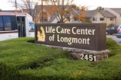 A roadside sign reading 'Life Care Center of Longmont' mounted on a low stone base surrounded by grass, with a bus, cars, and buildings in the background.