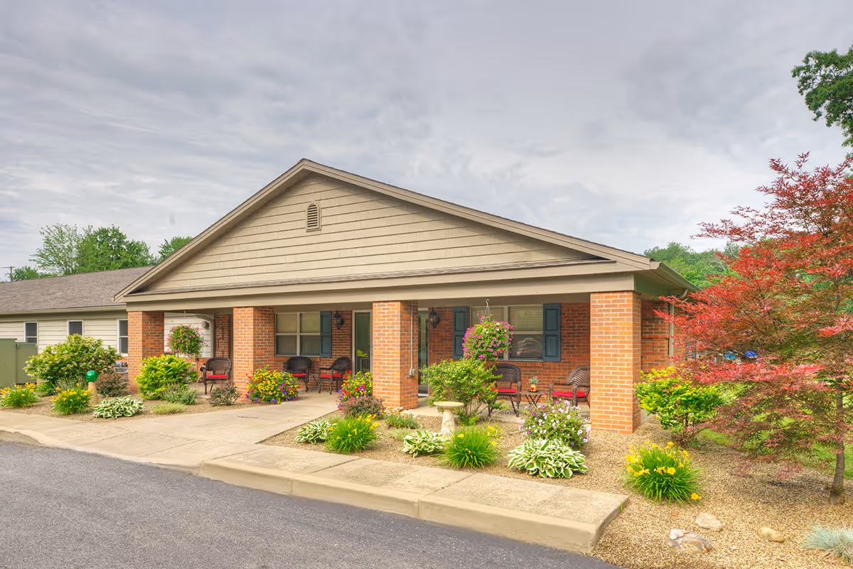 Front exterior view of Austintown Healthcare Center showing a single-story brick building with a covered porch. The porch has several chairs and hanging flower baskets. The surrounding area is landscaped with various shrubs, flowers, and a small tree with red leaves.