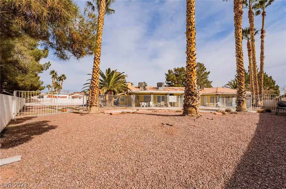 Outdoor area of a senior living facility with a gravel ground, several tall palm trees, and a white metal fence. In the background, there is a single-story building with a tiled roof and multiple windows and doors, some with white patio chairs outside. The sky is partly cloudy.
