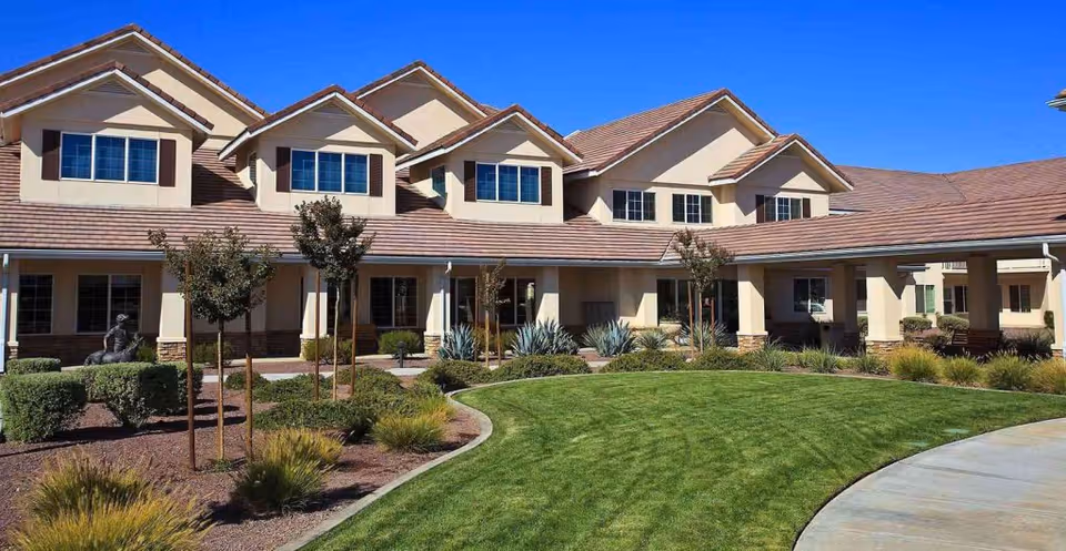 Exterior view of a senior living facility building with beige walls, multiple windows, and a tiled roof. The foreground features a well-maintained lawn, landscaped bushes, small trees, and a curved concrete walkway under a clear blue sky.