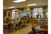 A spacious dining room with multiple tables and chairs arranged neatly. The room has large windows with blinds, potted plants on the windowsills, and a pool table in the corner. The ceiling has recessed lighting, and the walls are decorated with framed pictures and wall hangings.