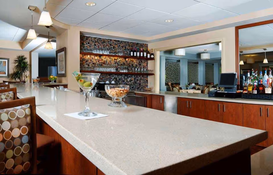 Interior view of a bar area with a long light-colored countertop, several bar stools with patterned cushions, a martini glass with olives, a bowl of snacks, shelves with bottles and glasses, and a large mirror reflecting a dining area with tables and chairs.