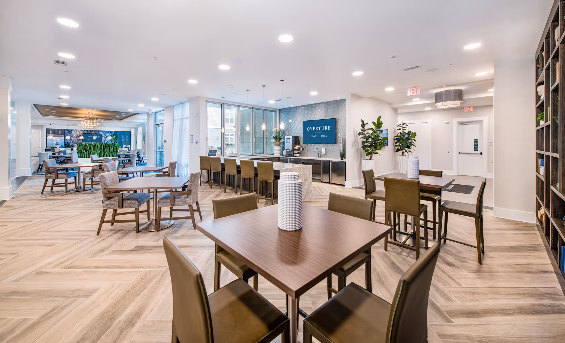 Bright modern communal dining area with tables, chairs, a long counter and an 'Overture Chapel Hill' sign on the back wall.