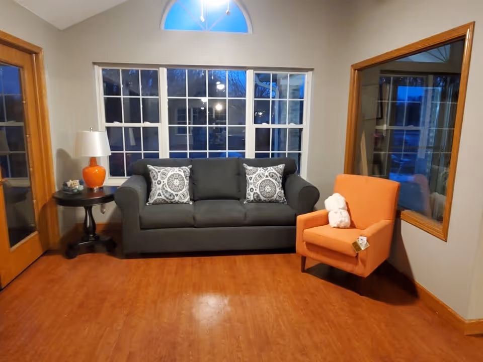 A cozy living room with a dark gray sofa adorned with two patterned pillows, an orange armchair with a white stuffed animal on it, a round wooden side table with an orange lamp, and large windows with white frames letting in evening light.