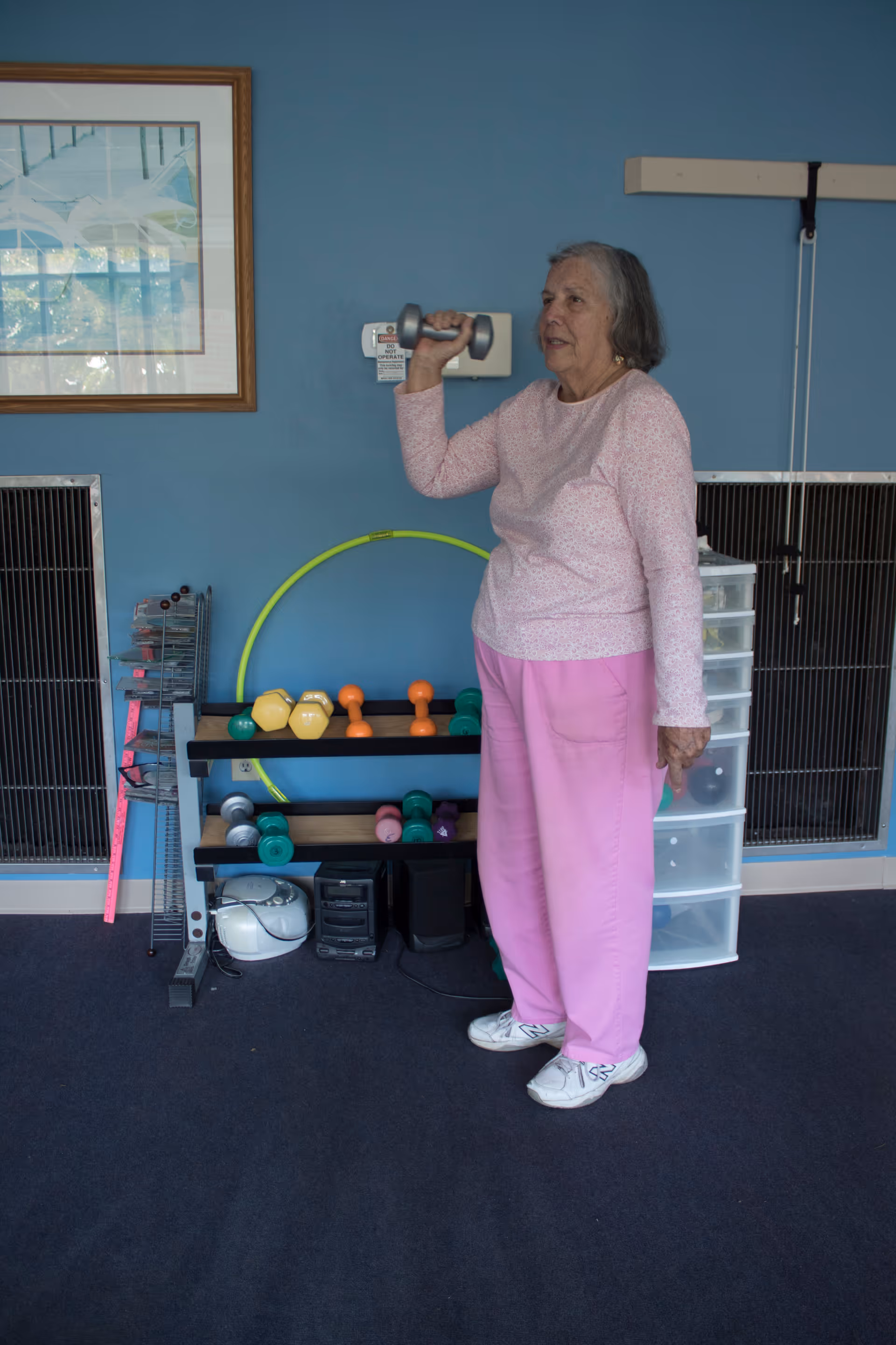An elderly woman wearing a pink long-sleeve shirt and pink pants is standing indoors holding a small dumbbell in her right hand, appearing to exercise. Behind her is a blue wall with a framed picture, a rack with various colored dumbbells, a yellow hula hoop, and a plastic drawer unit.