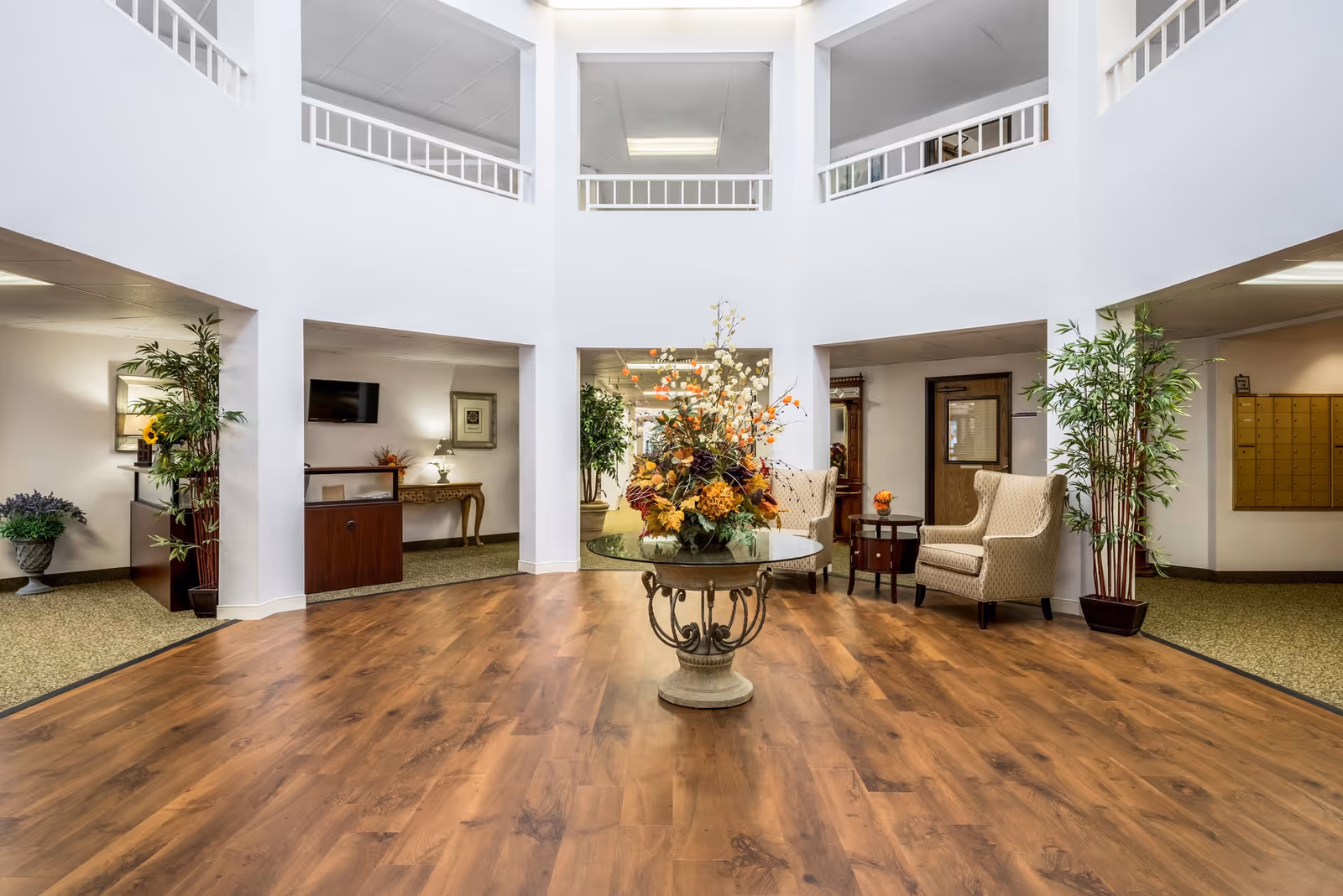Bright and spacious senior living facility lobby with a round table in the center holding a large floral arrangement. The room features wood flooring in the middle and carpeted areas around the edges. There are two beige upholstered armchairs with a small round table between them on the right side, potted plants on either side, and a reception desk with a TV mounted on the wall in the background.