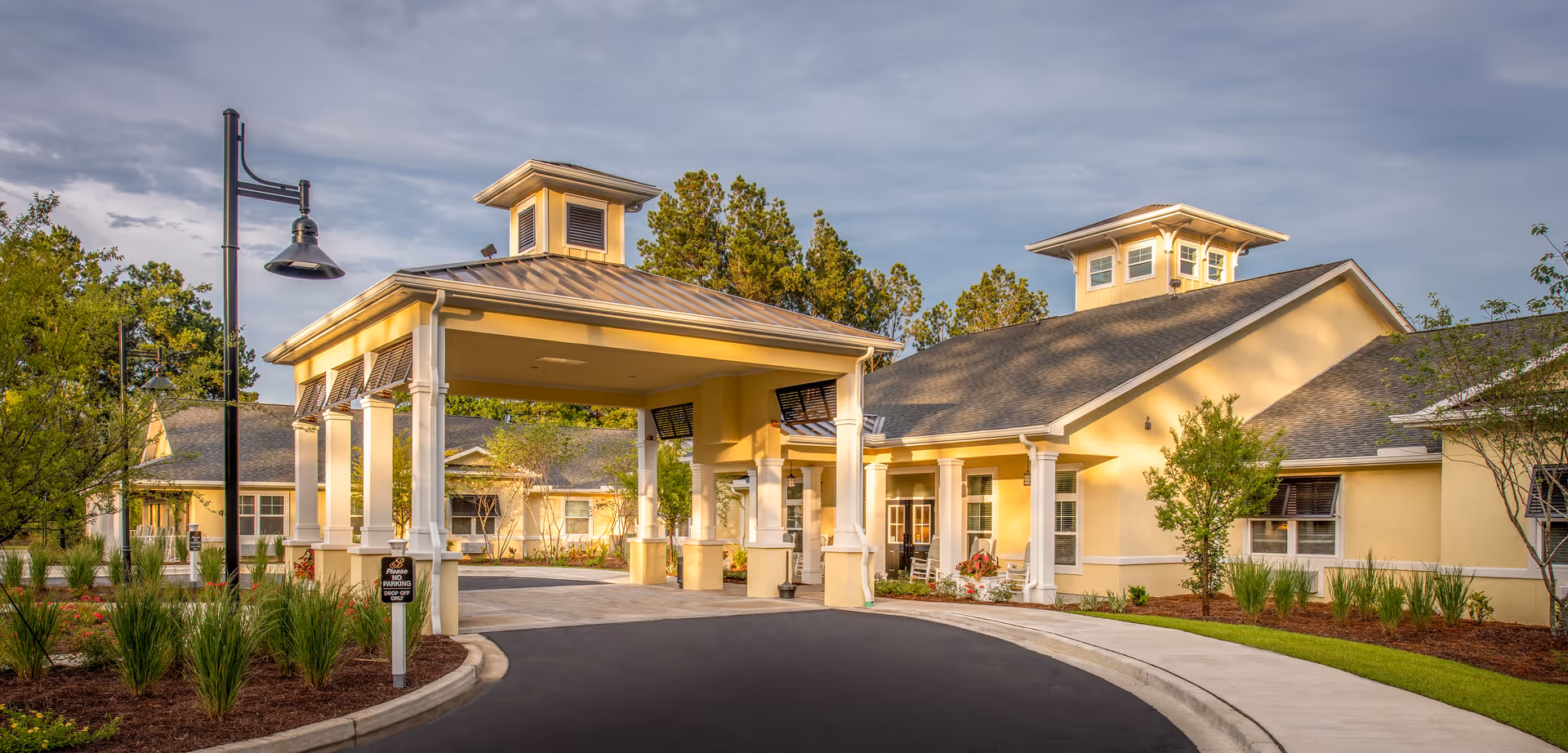 Covered porte-cochère entrance of a yellow single-story senior living facility with landscaped driveway.