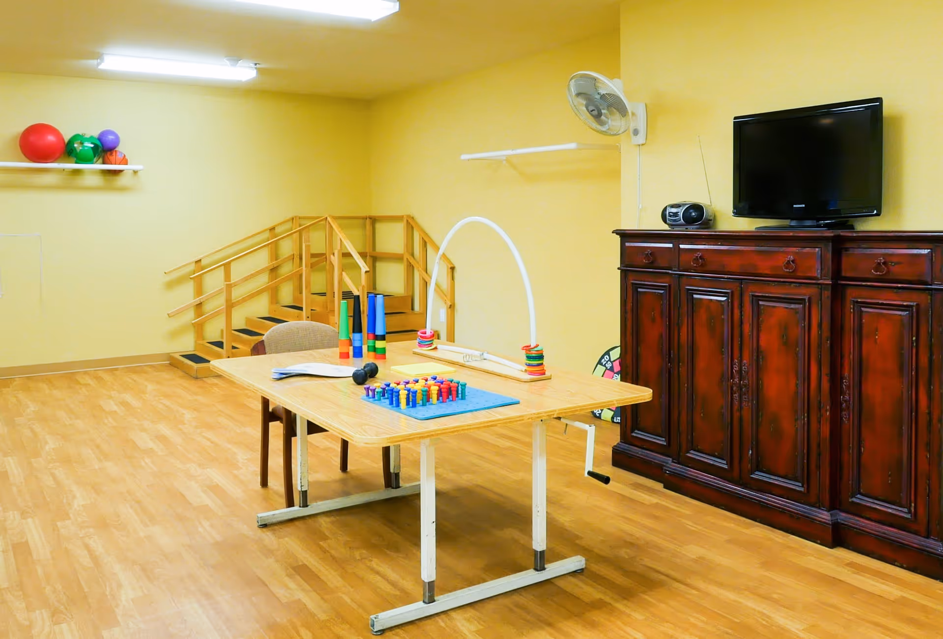 A therapy or activity room with a wooden table holding various therapy tools and games, a chair, a wooden cabinet with a TV and radio on top, a wall-mounted fan, and a small set of stairs with handrails in the background. The walls are painted yellow and the floor has a wood finish.