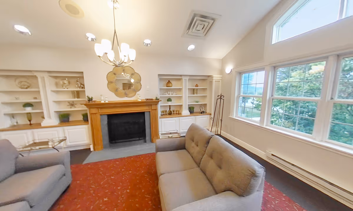 Well-lit common living room with sofas facing a fireplace, built-in shelving, large windows and a chandelier.