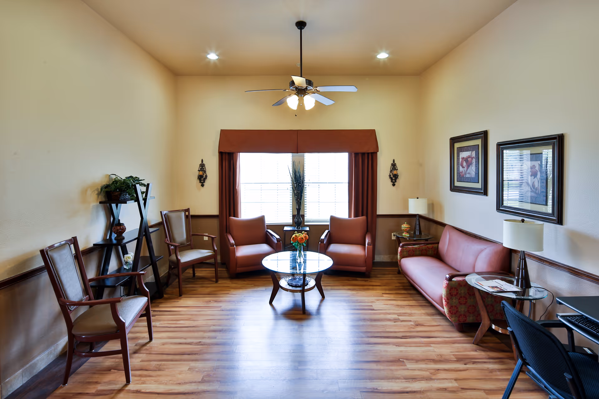 Sunlit communal living room with armchairs and a sofa arranged around a glass coffee table beneath a ceiling fan.