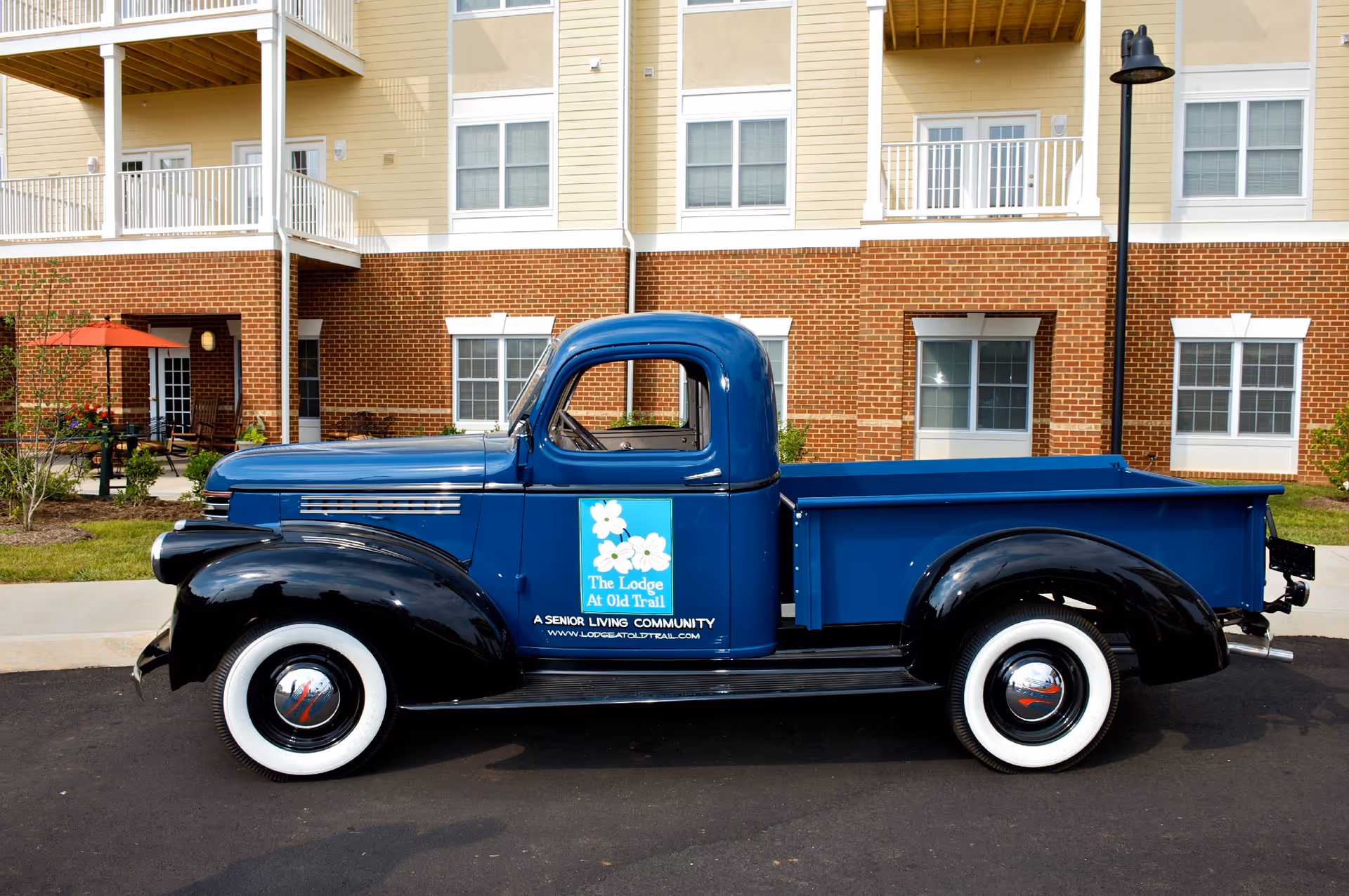 A vintage blue and black pickup truck with whitewall tires parked in front of a senior living community building. The truck has a logo on the door that reads 'The Lodge At Old Trail, A Senior Living Community' with a website URL. The building behind has brick and beige siding with multiple windows and balconies.