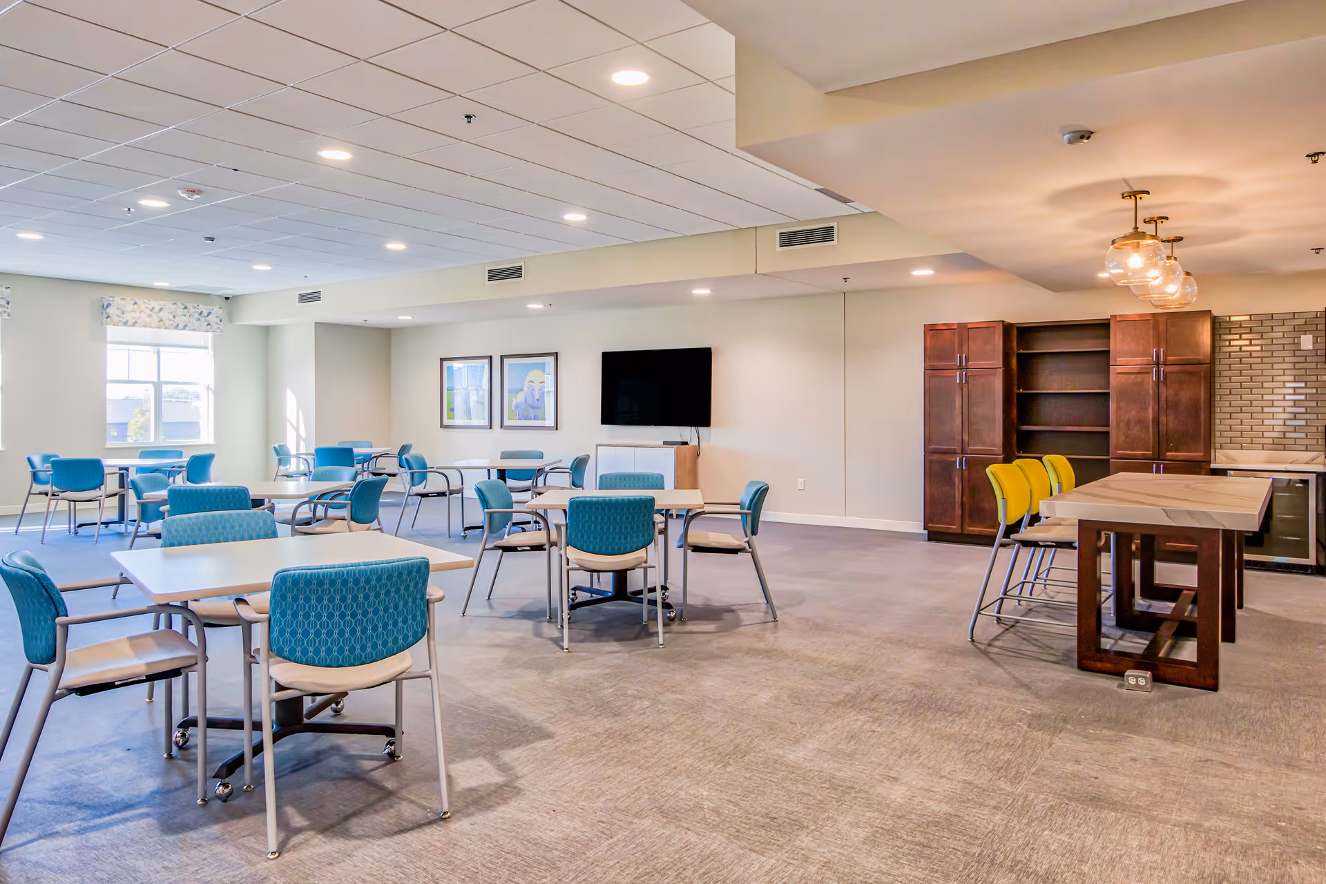 A spacious senior living dining area with multiple tables and blue chairs arranged neatly. There is a large flat-screen TV mounted on the wall, two framed pictures beside it, and a window letting in natural light. On the right side, there is a kitchen area with wooden cabinets, a countertop, and yellow bar stools under hanging light fixtures.