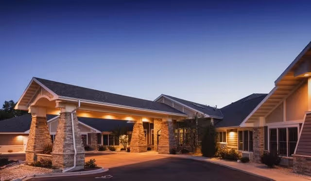 Exterior view of a senior living facility building at dusk with warm lighting illuminating the entrance area supported by stone pillars and a driveway in front.