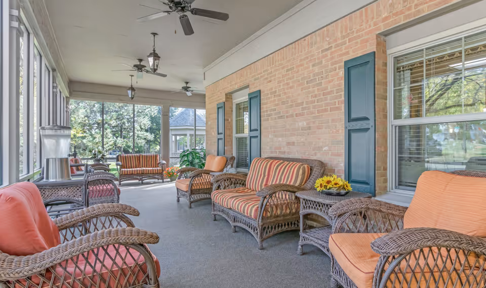 A screened-in porch area with wicker furniture including chairs and loveseats with orange and striped cushions. The porch has ceiling fans and lantern-style light fixtures, with a brick wall and windows on one side and large screened windows looking out to a green outdoor area.
