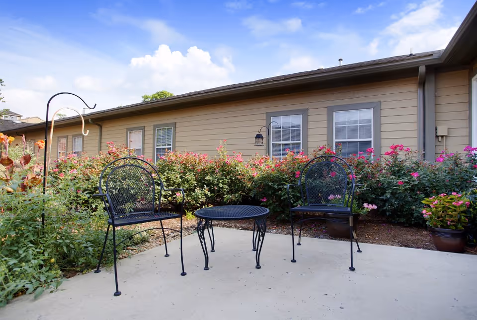 Outdoor patio area with two black metal chairs and a matching round table on a concrete surface, surrounded by blooming pink flowers and green shrubs, with a beige building and windows in the background under a partly cloudy blue sky.