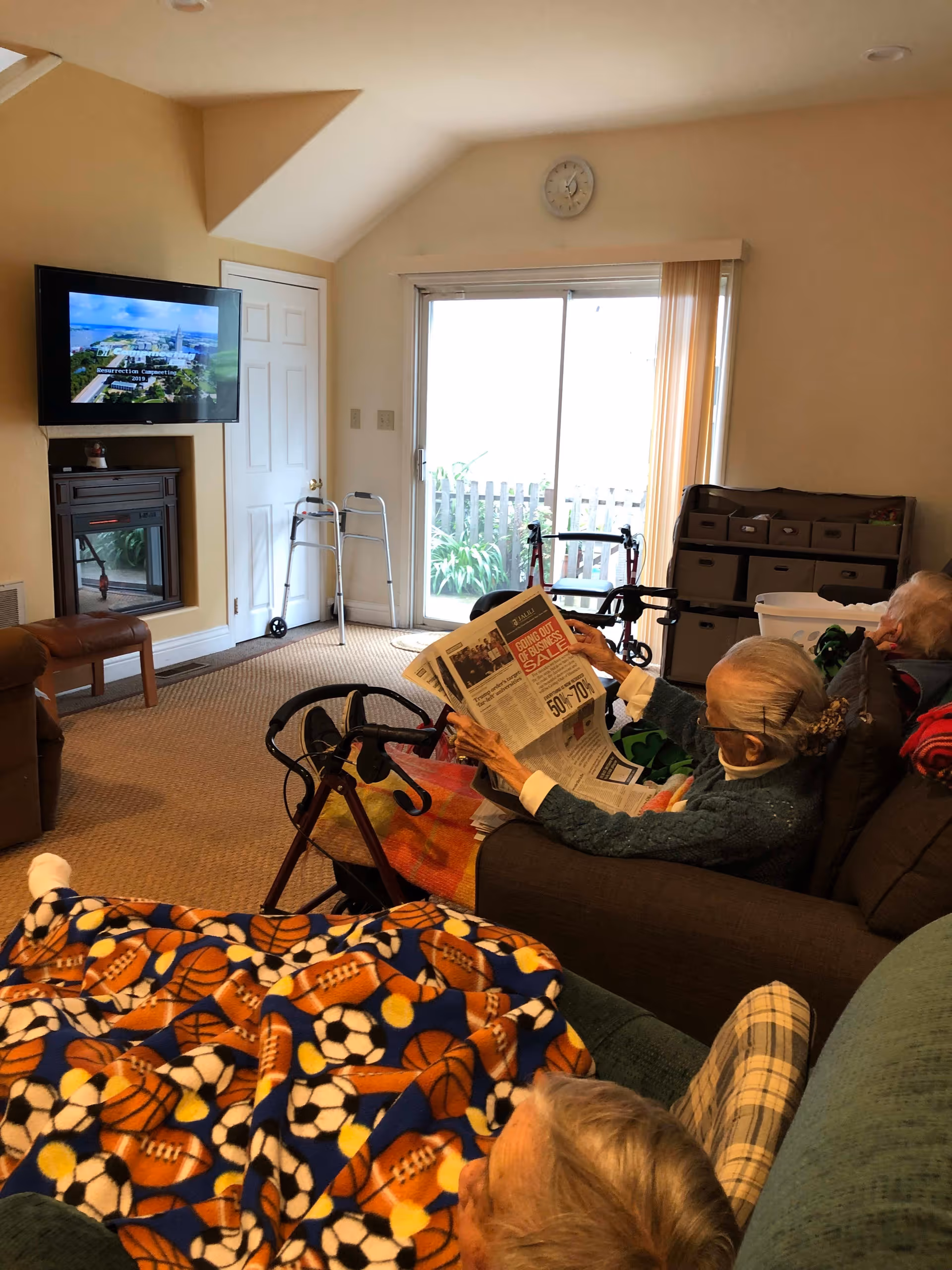 A cozy living room in a senior living facility with two elderly women seated on couches. One woman is reading a newspaper while the other is resting under a colorful sports-themed blanket. The room has a wall-mounted TV, a walker near a closed door, a sliding glass door with vertical blinds, and a storage unit with drawers. The carpeted floor and soft lighting create a warm atmosphere.
