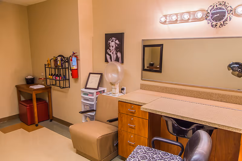 Interior of a salon or beauty treatment room with a beige chair, a countertop with drawers, a large mirror with lights above it, a hair dryer, and a black and white photo of Marilyn Monroe on the wall. There is also a small table with a red bin underneath and a wall-mounted rack holding various hair products and a fire extinguisher.