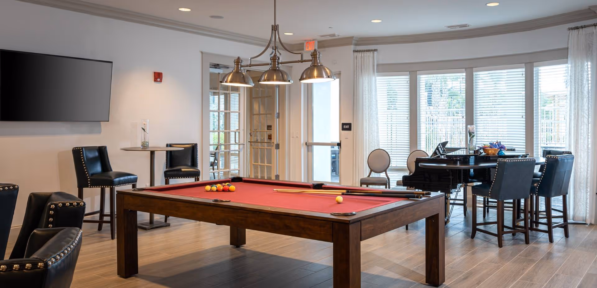 A well-lit recreational room with a red felt pool table in the center, surrounded by black leather chairs with nailhead trim. There is a round table with chairs near large windows with white blinds, and a flat-screen TV mounted on the wall. The room has light-colored walls and wood flooring, with a modern light fixture hanging above the pool table.