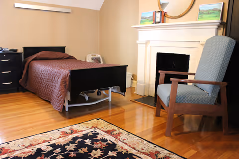 A bedroom with a single bed covered with a brown quilt, a wooden nightstand with a telephone, a fireplace with a round mirror and paintings above it, a wooden armchair with blue upholstery, and a patterned area rug on a wooden floor.