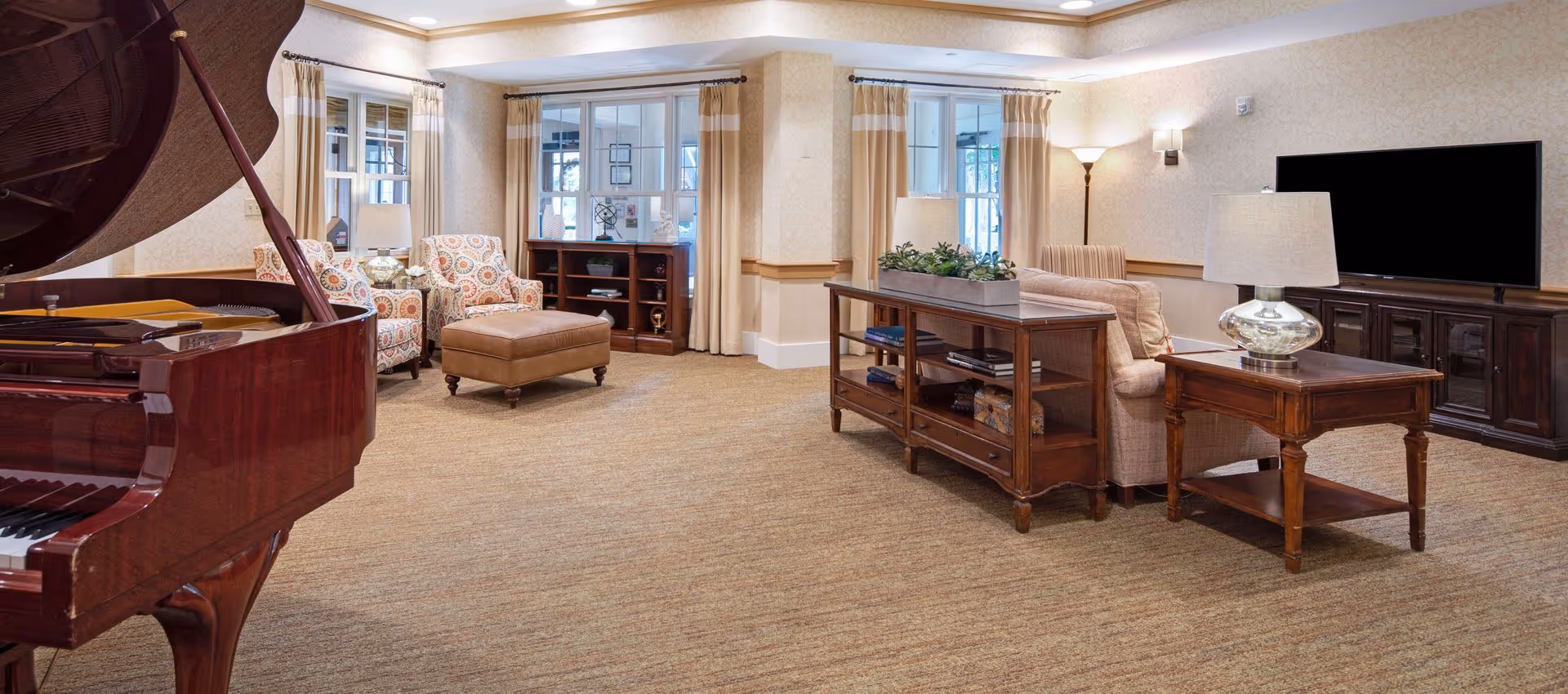 A spacious communal living room with a grand piano, upholstered chairs and ottoman, wooden tables and shelving, and a wall-mounted TV.