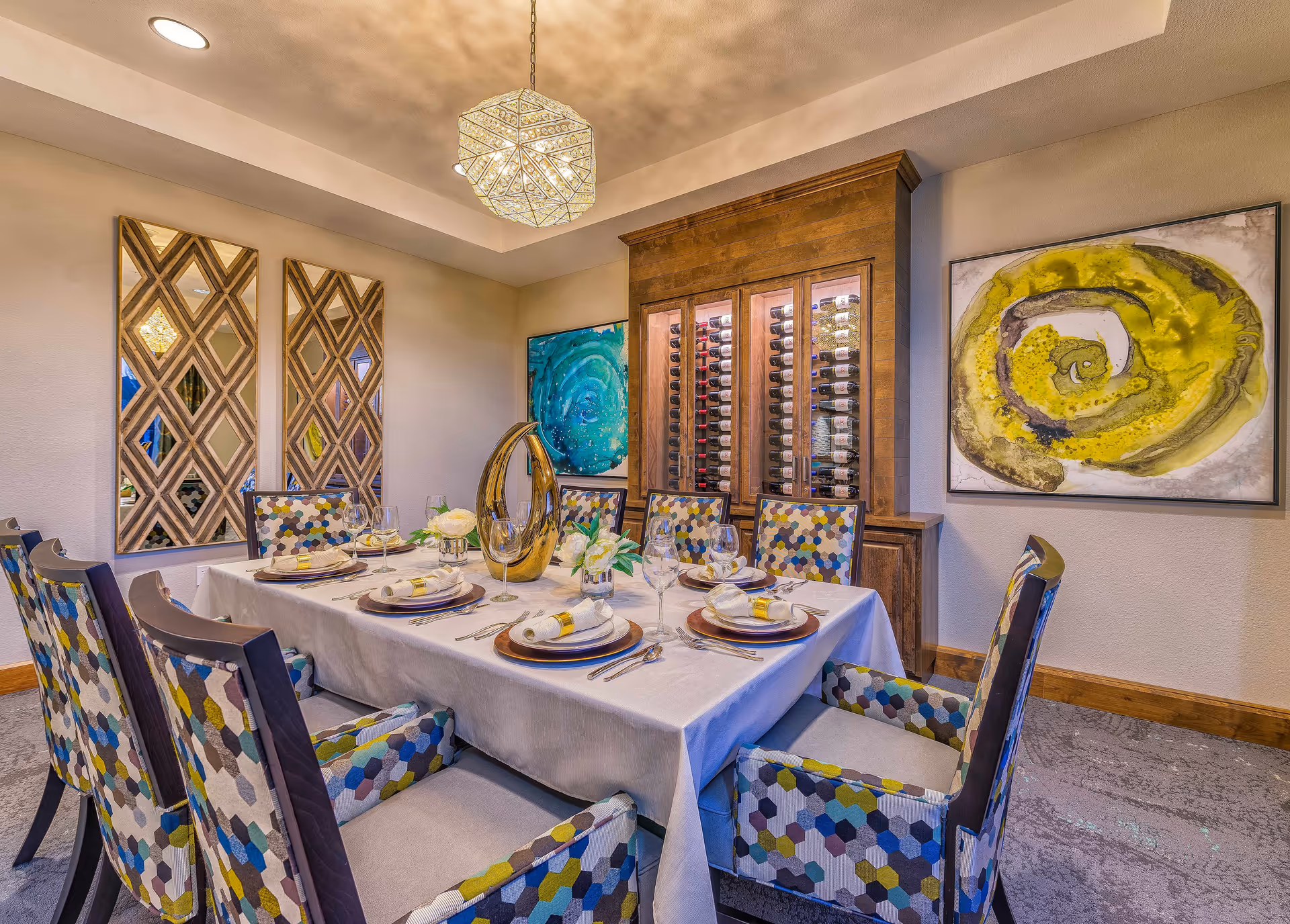 A dining room with a rectangular table covered with a white tablecloth, set for six with plates, napkins, wine glasses, and cutlery. The chairs have colorful hexagonal patterned upholstery. The room features a wooden wine rack filled with bottles, two large abstract paintings on the walls, and two decorative mirrors. A geometric pendant light hangs from the ceiling.