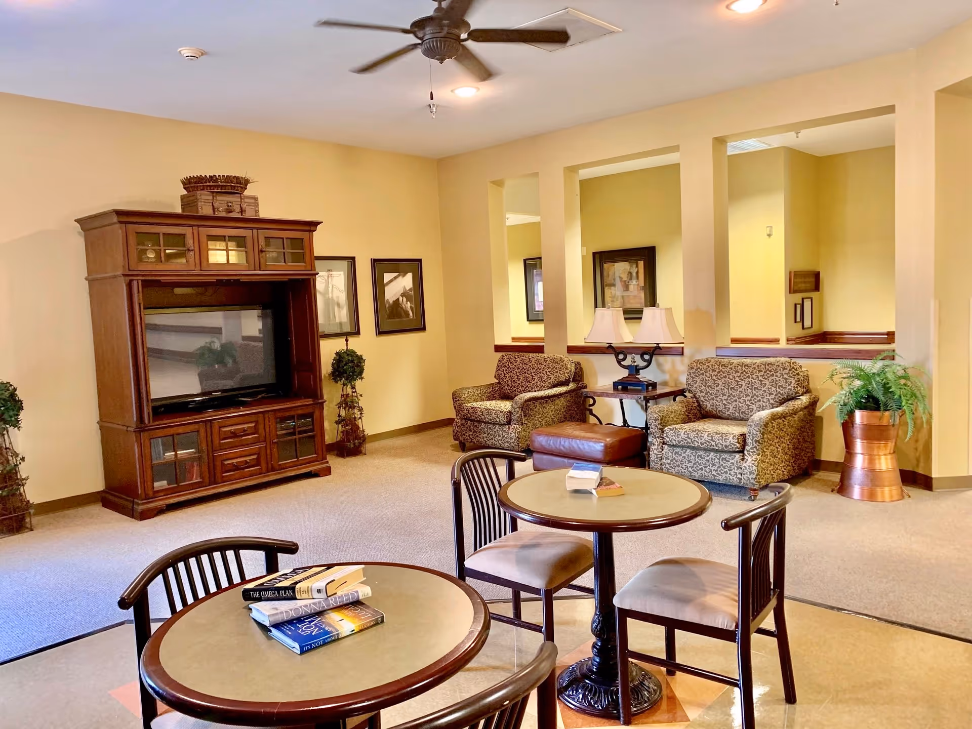 A cozy living room area in a senior living facility featuring a large wooden entertainment center with a TV, two patterned armchairs with a brown ottoman between them, a side table with a lamp, and two round tables each with two chairs. There are framed pictures on the walls and potted plants adding greenery to the space.