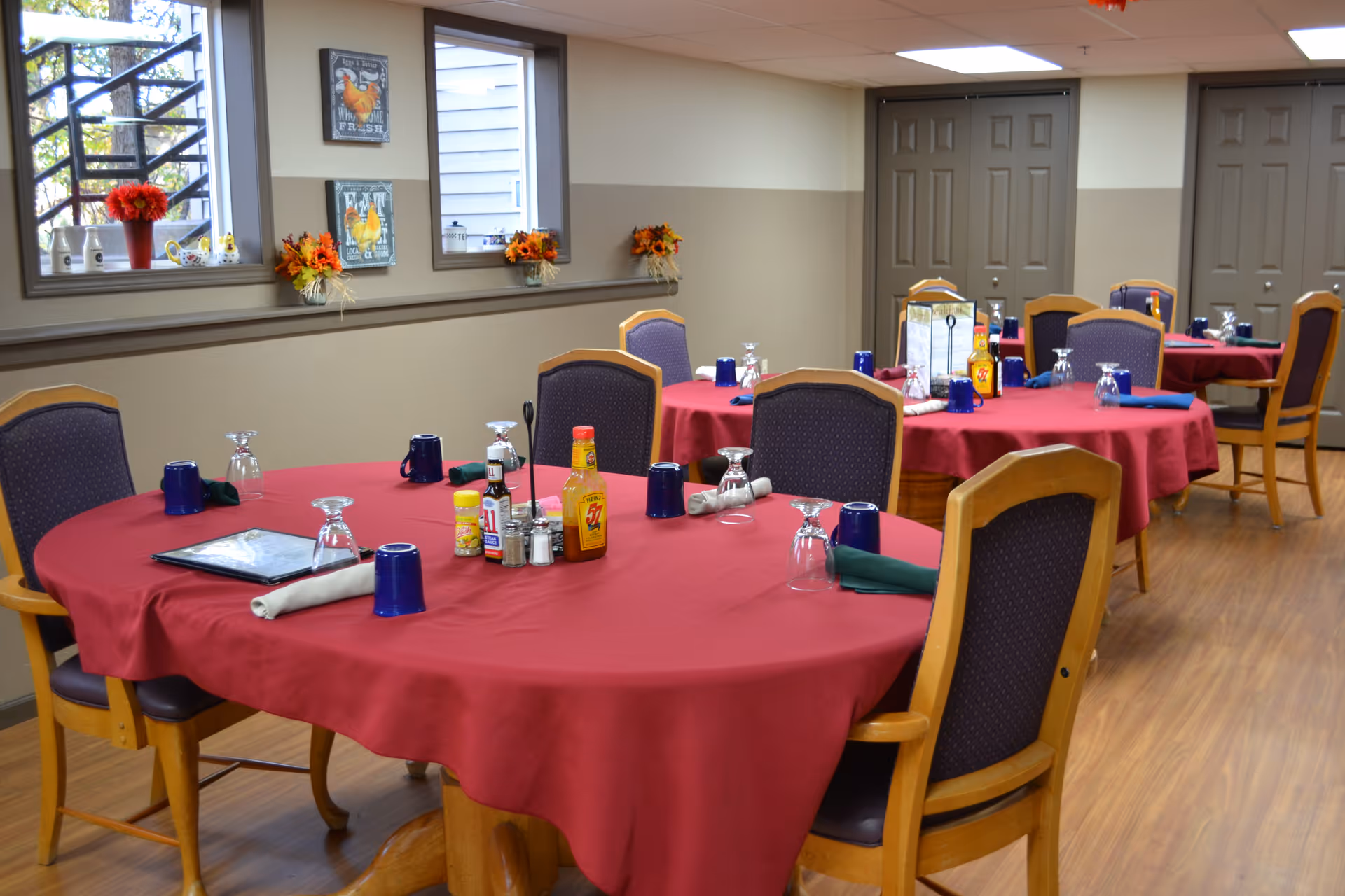 Communal dining room with round tables draped in red tablecloths, place settings, and wooden chairs.